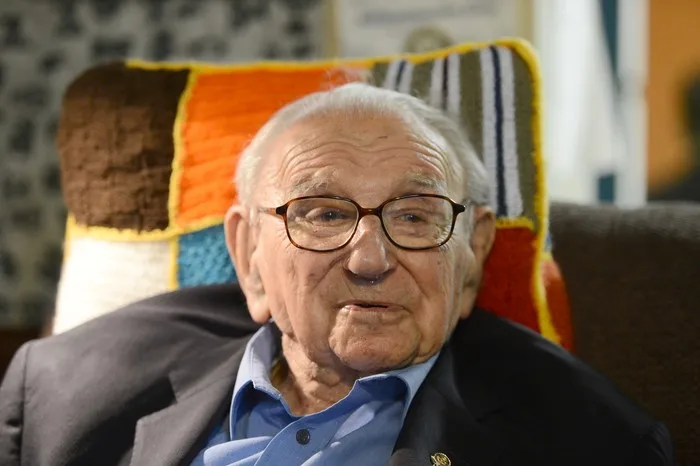 Sir Nicholas Winton wearing a suit and glasses. He is sat on a sofa