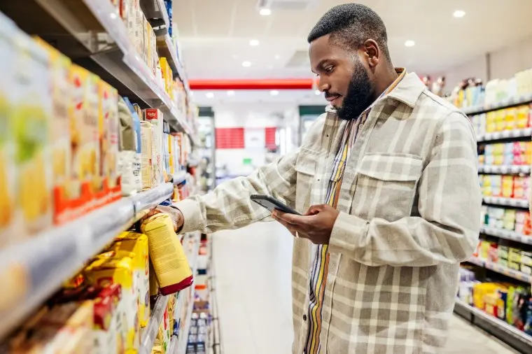 A man using his smartphone to look up ingredients and prices wile choosing items in his local supermarkets.