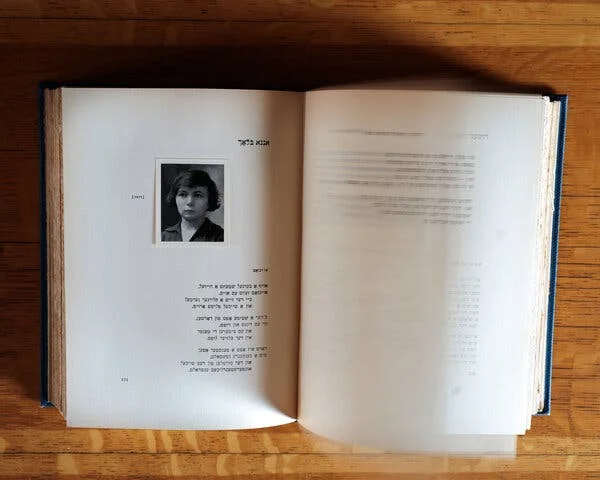 A book, atop a wooden table, opened to a Yiddish poem written by a woman who’s seen in a passport-size, black-and-white photo above the text.