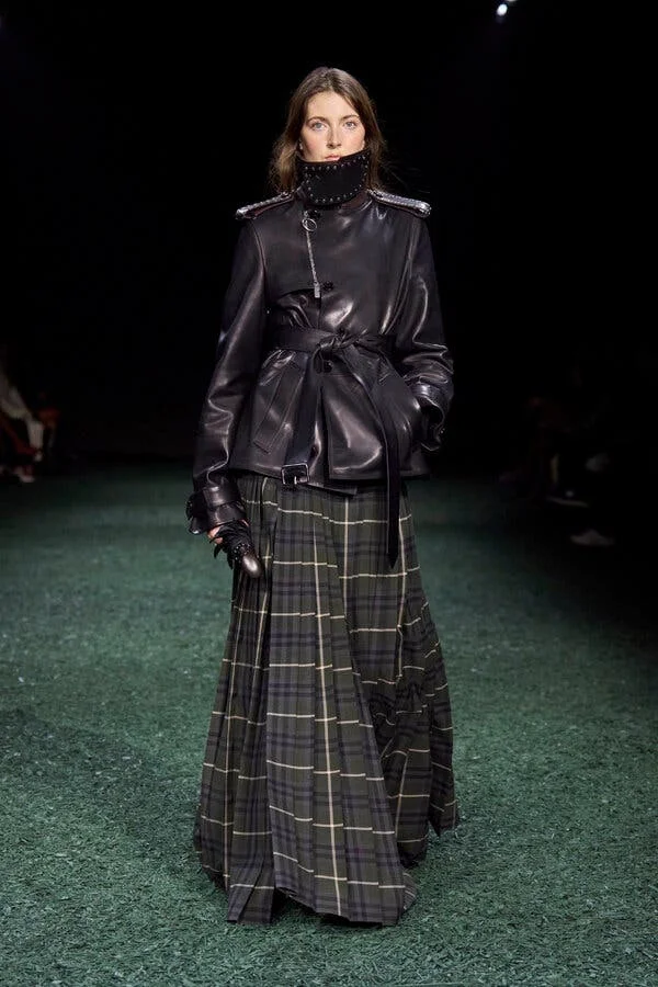 Maya Wigram stands on a grassy runway wearing a belted black leather jacket with a funnel collar and a floor-length dark-green plaid skirt.