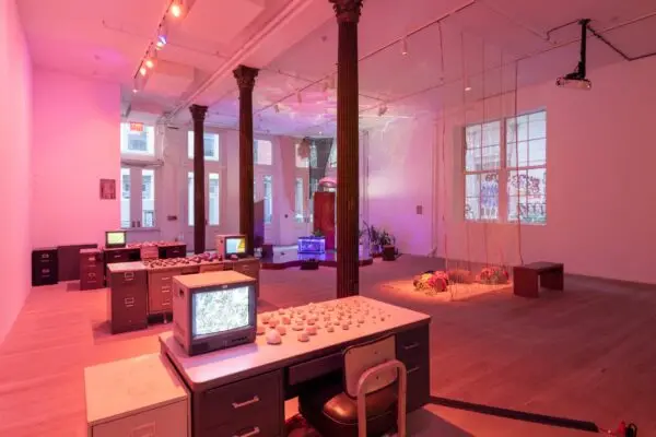An installation image of a pink hued room with various objects including desks, file cabinets, monitors, houseplants, and more.