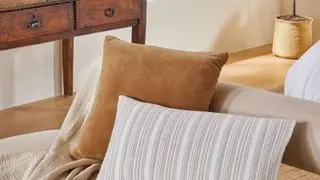 A cream bench with a brown square throw pillow and a rectangular white and gray striped throw pillow on it, with a vintage dark brown sideboard to the left of it