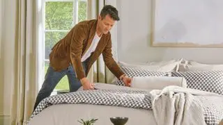 A picture of Nate Berkus, a man wearing a brown suede blazer, white t-shirt and jeans, making a beige bed with black and white checked trim, with a window with long beige curtains behind him