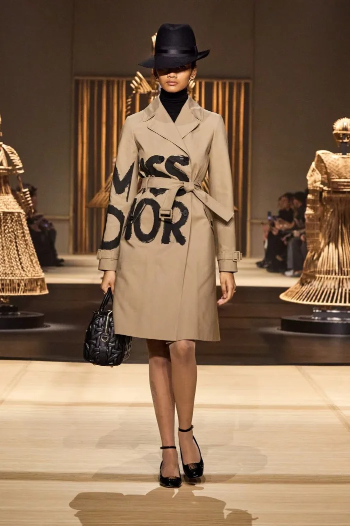 A model wears a trenchcoat emblazoned with the words Miss Dior