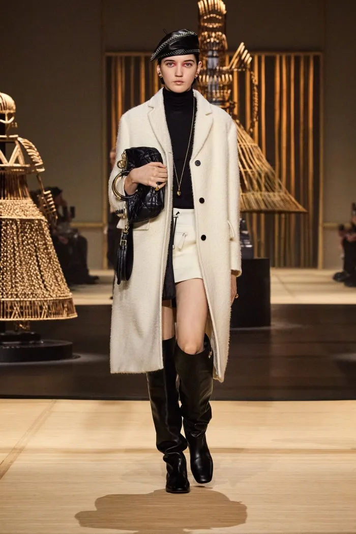 A model wears a long cream coat over cream miniskirt and black polo neck sweater. She also wears knee high boots and black beret