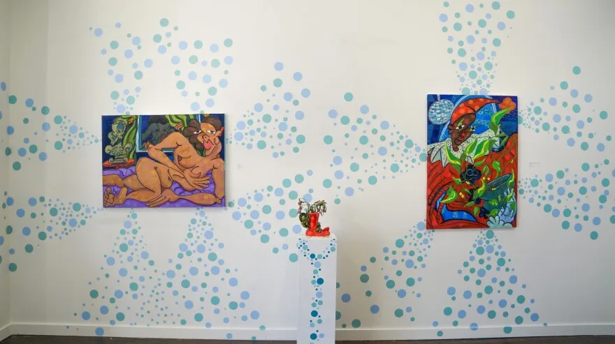 Two paintings and a brightly colored vase on display against a wall and platform that is white with blue spots of various sizes painted on it.
