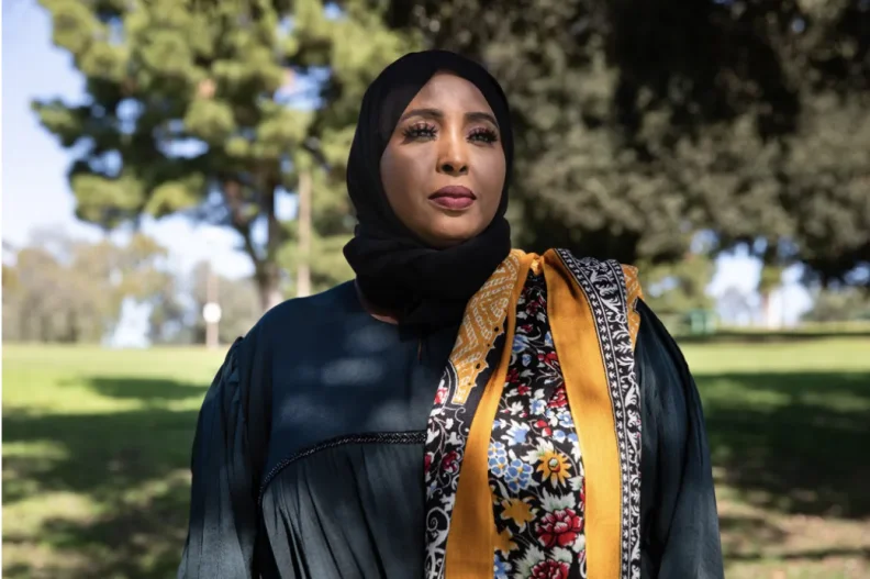 A woman with brown skin tone wears a hijab, brightly colored scarf and a dark shirt. She gazes off to the right of the viewer. It's sunny out, with just a few trees casting shadows over the grass behind her.