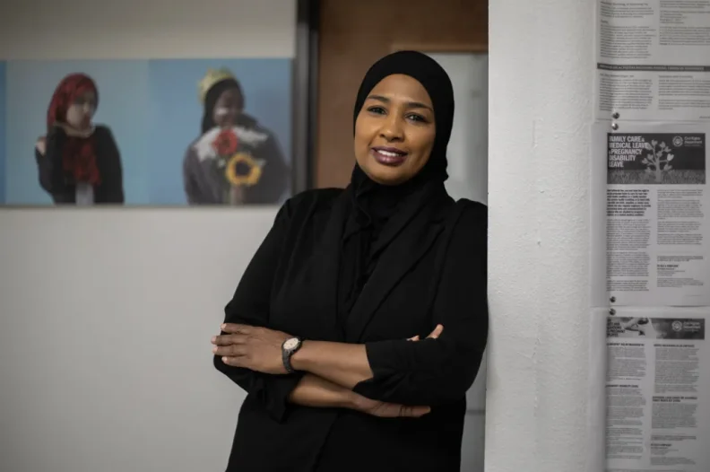 A woman with brown skin wears a black hijab and outfit as she leans against a doorway, smiling at the camera. Behind her, blurred in the background, are small paintings on the wall of other young women, wearing hijabs. One has flowers in front of her.