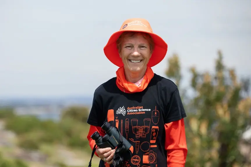 Annie Lane wears a black and orange shirt, hat and holds binoculars.