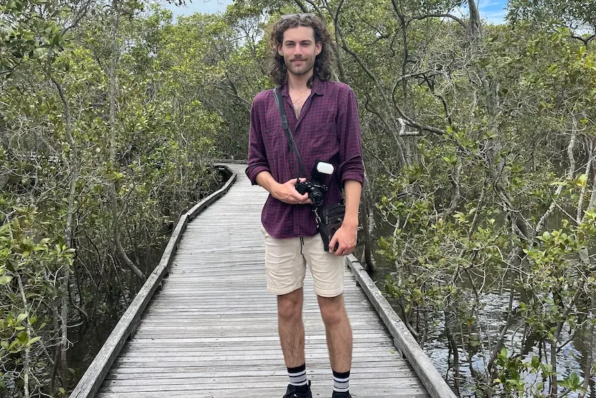 A young man stands on a wooden boardwalk with trees in the background.