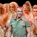 A man in a short-sleeved shirt beams as models with blond hair stand behind him.