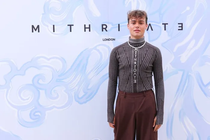 Bradley Riches in a long-sleeved grey shirt and brown trousers. He is posing front of a light blue background