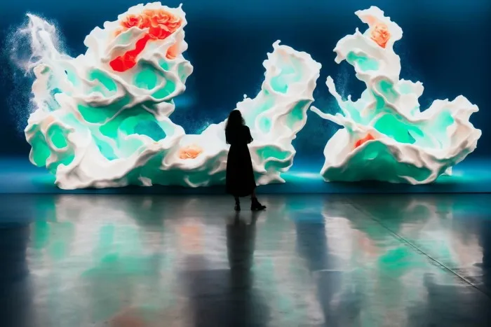 A woman in an art gallery is dwarfed by large images of coral-like structures projected on screens behind her