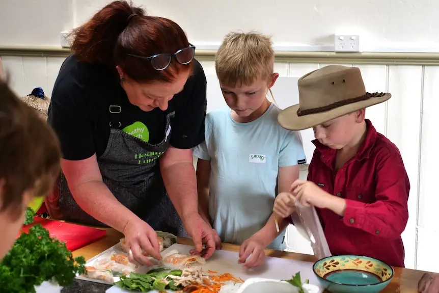 Two boys make rice paper rolls with the help of a women.