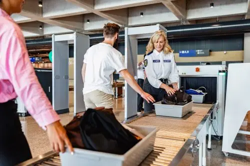 Two travelers putting their bags on the belt at airport security while a female TSA agent helps them.