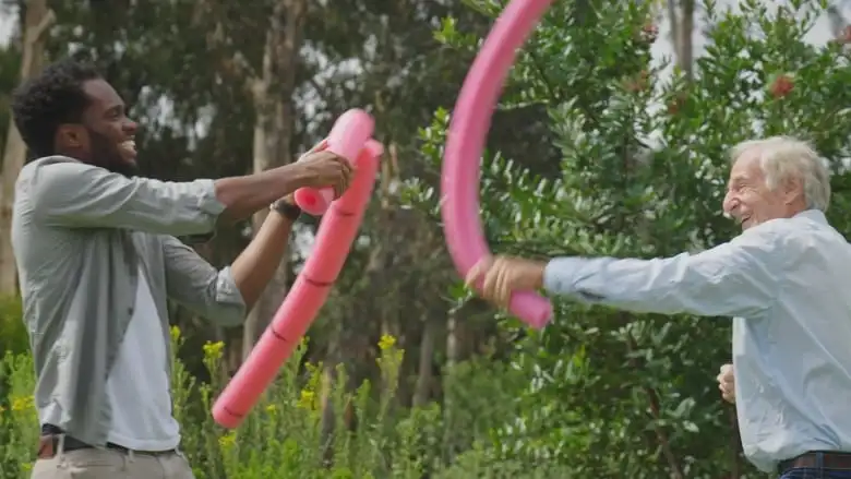 Two men flight playfully with pink pool noodles. One noodle is thicker than the other.