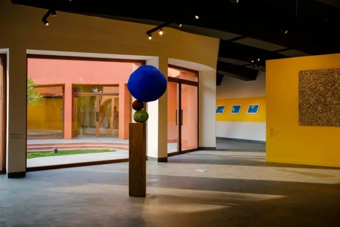 An art gallery contains paintings and sculptures, including a column with rough spheres of various sizes balanced on it; at the top is a large vivid blue orb