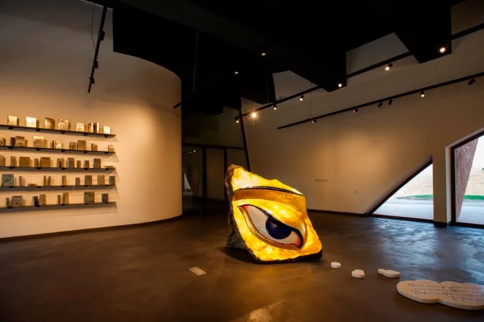 An art gallery features artworks including a set of shelves with golden packets and a large sculpture of a gold-decorated eye with a vivid blue iris