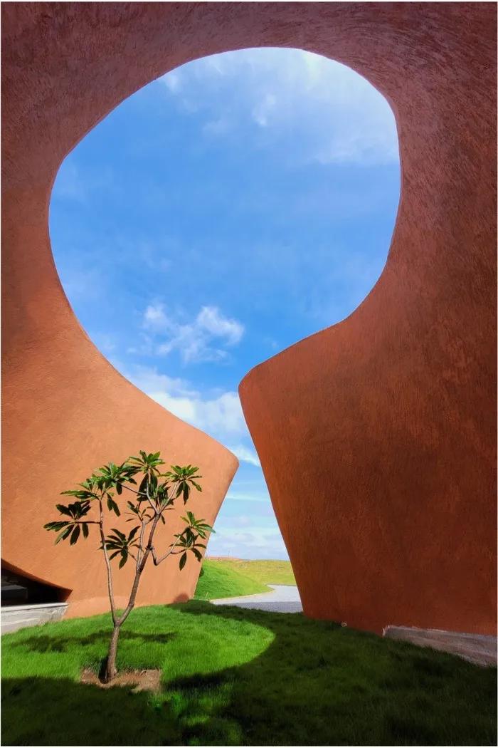 A section of a reddish building forms a keyhole-like shape, through which blue sky can be seen; in the foreground grows a small tree