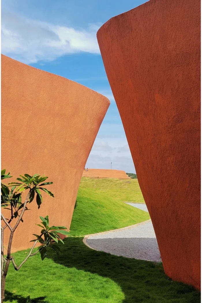 Two parts of a curved reddish complex of buildings lean towards each other; around them is grass and vegetation