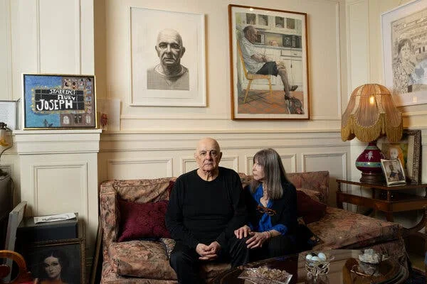 A man and a woman sit on a sofa in an apartment with art on the walls.