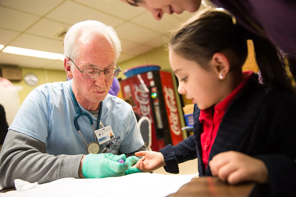 a doctor takes a blood sample from a small girl with pale skin and dark hair in a school cafeteria