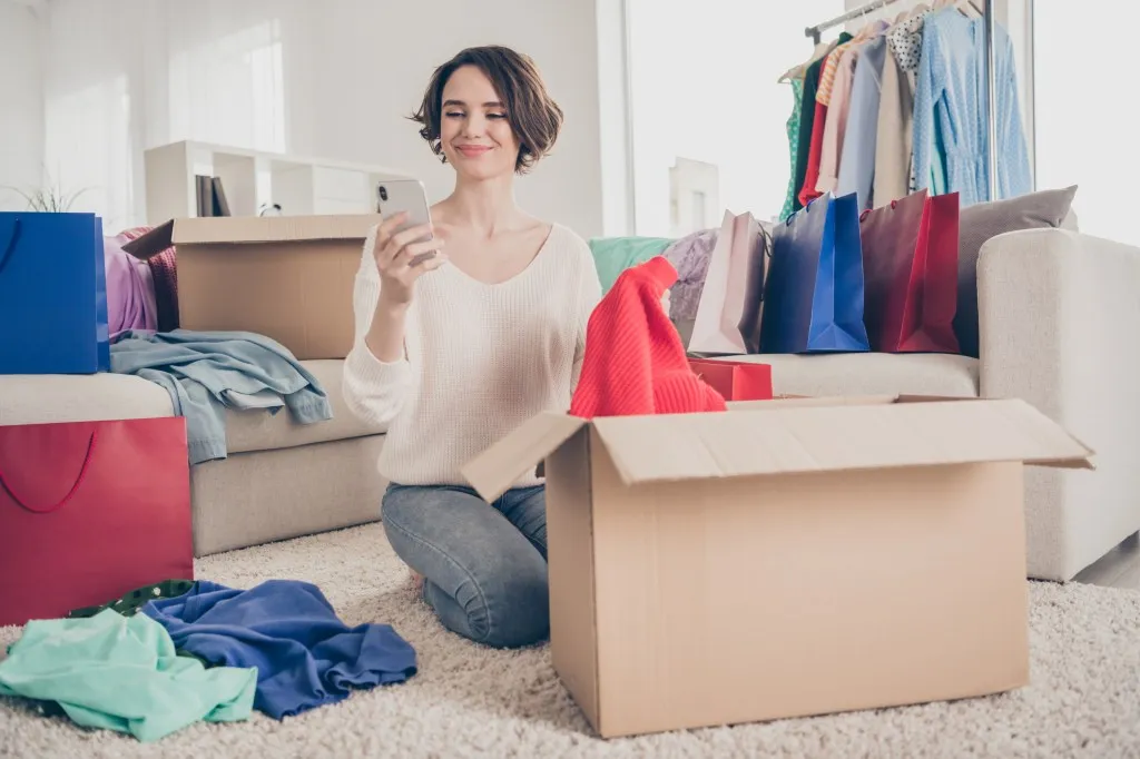 Full size photo of cheerful young lady sit floor look phone donate clothes boxes apartment smile indoors in living room.