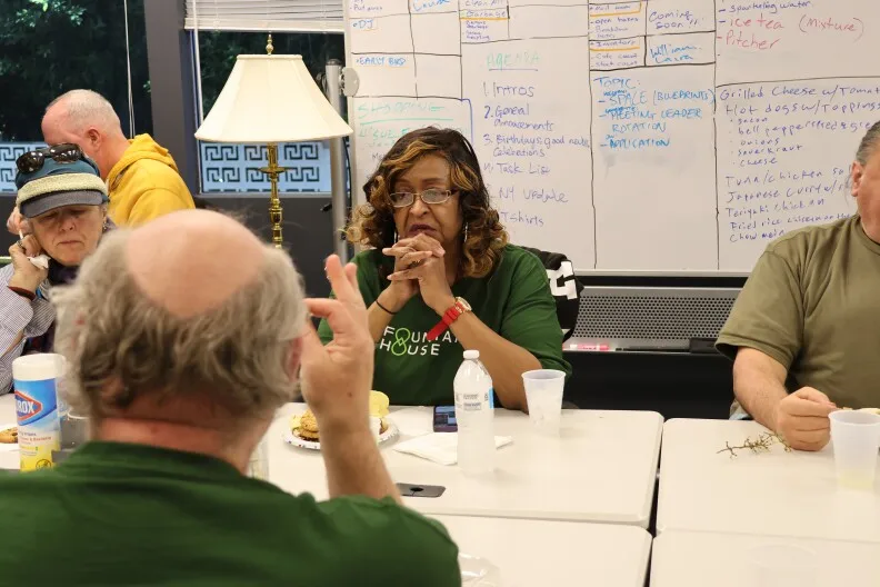 Georgette Darby sits at a conference table table with her hands folded. Behind her is a white board various writing on it. 