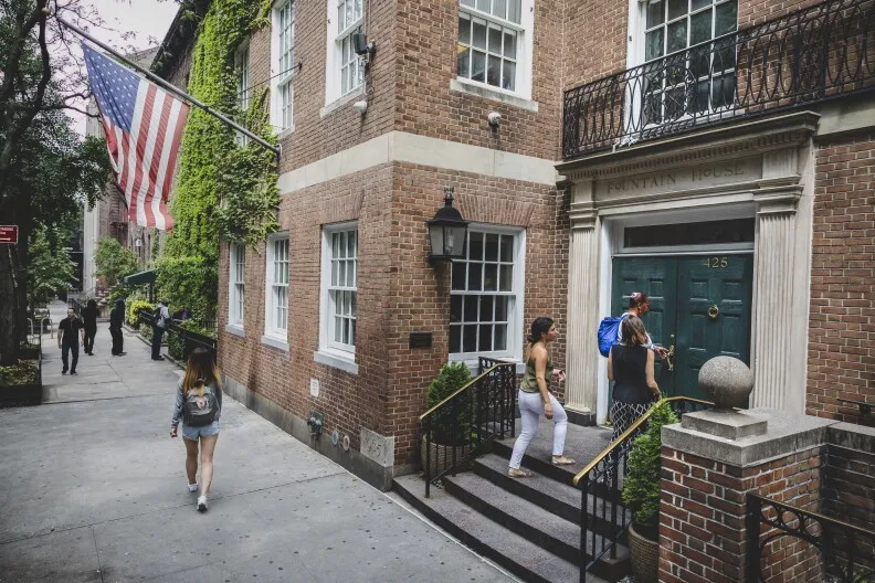 An exterior photo of Fountain House's main clubhouse in New York City. A distinguished looking brick building stands on a New York street. A large American flag hangs out front. Two people walk up the front steps to enter the building. Above the threshold reads 'Fountain House.'