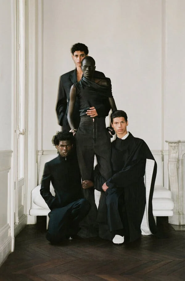 Four models, all dressed in black, with two seated, and two standing. The model standing in front has his legs held by the two seated models, and a model standing behind him, with his hand around his waist.