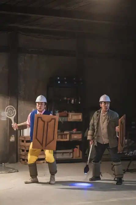 Still shows the same two fellows as previously, now wearing white hard hats and holding makeshift shields and what looks like electric bug-killing tennis rackets