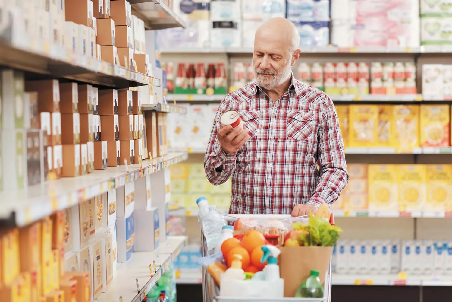 photo of a man in a supermarket standing with a full cart, holding a can and reading the label