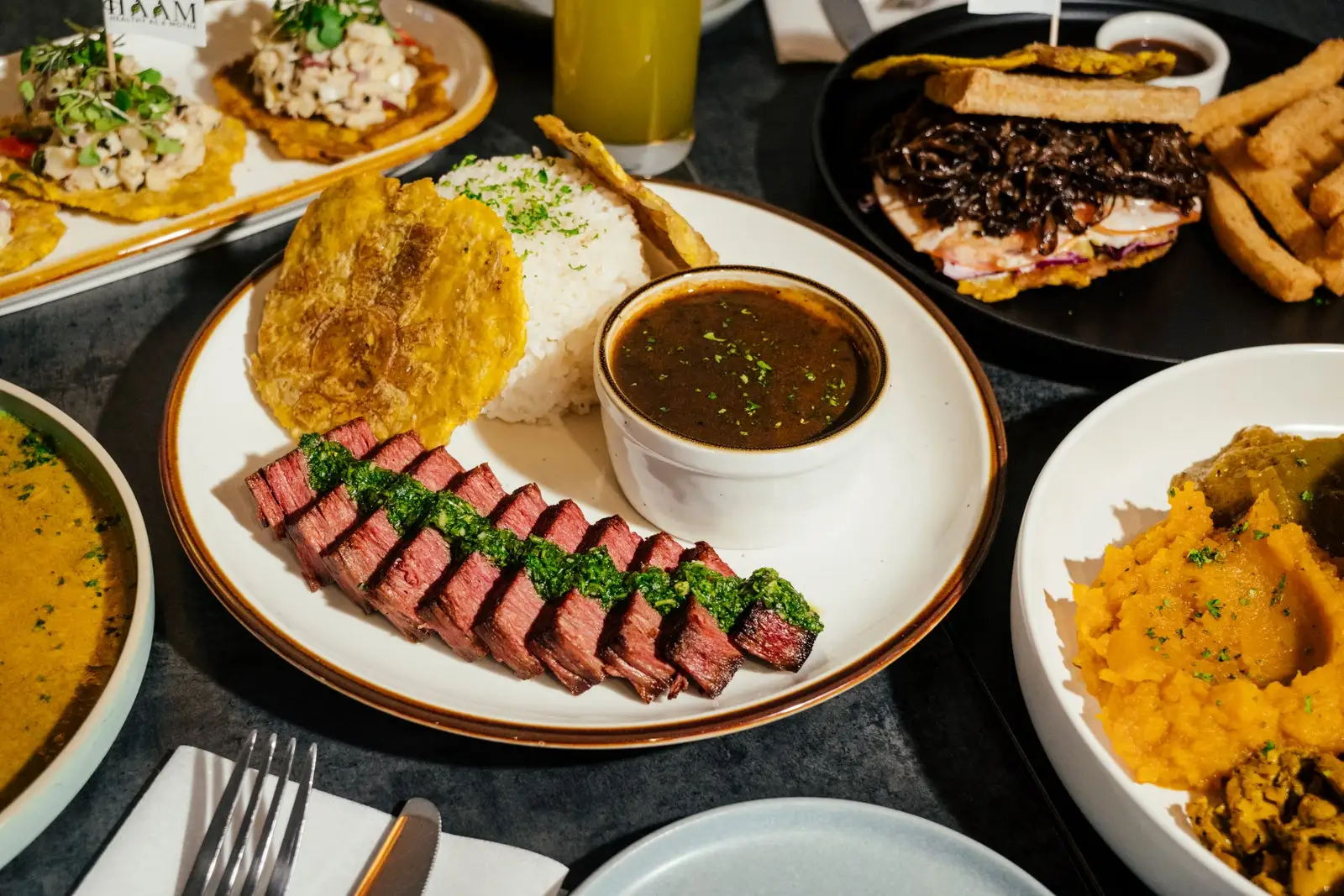 Slices of vegan steak drizzled with chimichurri and served with white rice tostones and black beans.