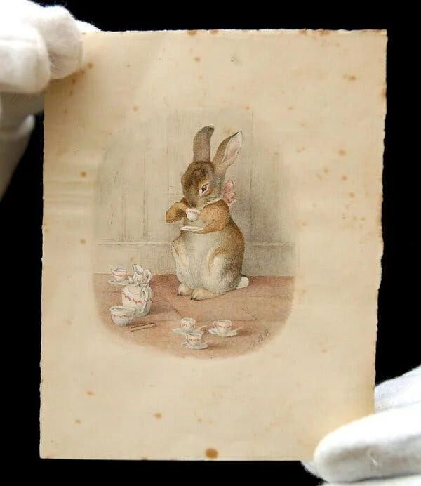 A color photograph shows white-gloved hands holding a Beatrix Potter illustration of a bunny with a pink bow around its neck standing on its hind legs and drinking a cup of tea from a tiny, white, pink-rimmed china cup. On the floor in front of the bunny is the rest of the tea set: a teapot, four more tiny cups and saucers, a small dairy pitcher and a sugar bowl.