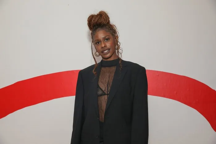 Zeze Millz in a black outfit stood in front of a white and red background, smiling. Her hair is up in a bun