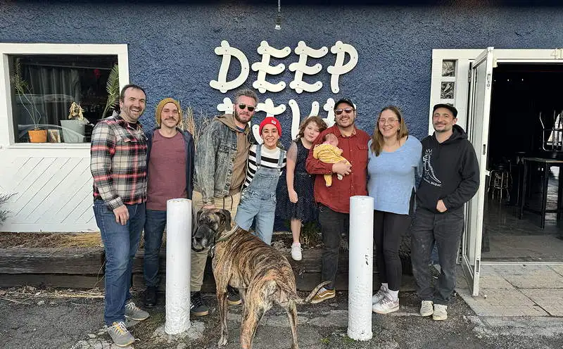 A row of people and a dog stand in front of a building with a sign that says Deep Dive. One of the people holds a baby.