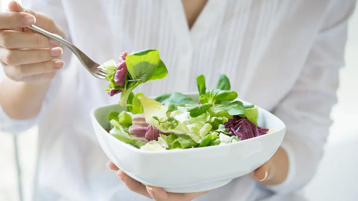 Close up shot of a woman holding a plate of fresh green salad in the beautiful morning light. She's holding a fork and she's about to eat the vegetarian food. Healthy eating and diet concept. Shallow depth of field with focus on the fork.