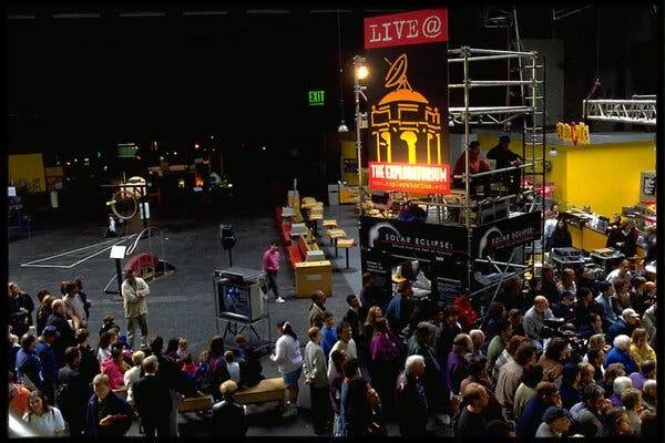 An audience in a studio. A large red sign says “Live @.”