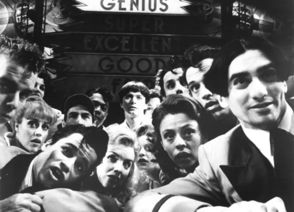 This black and white close-up image shows the 1993 cast of ‘Tommy,’ dressed in 60s mod clothing, crowded around a pinball machine, staring with fascination into the camera. Michael Cerveris stands center as Tommy, dressed in white, with a dark bowl cut and a blank expression. The scoreboard behind them has lit up the word “Genius.”