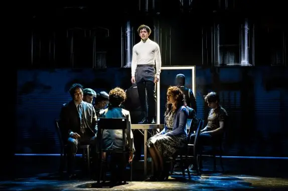 Tommy, played by a Moroccan American actor with dark curly hair, stands on top of a dining table during a Christmas dinner scene. He wears a bright white turtleneck and black pants. Young Tommy sits at the table directly in front of him, with the rest of his family on either side. A full-length mirror with a white, glowing frame stands behind them.