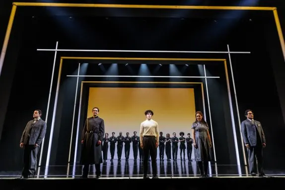 The stage is framed in yellow and white light, with a yellow-to-white gradient backdrop. The ensemble, dressed in all black, stands in a row upstage with hands clasped. At the front of the stage, Uncle Ernie, Cousin Kevin, Tommy, Mrs. Walker, and Captain Walker stand facing the audience. Uncle Ernie is a middle-aged white man with a dark combover, wearing a grey suit. Cousin Kevin is a white young adult with dark hair pushed back, wearing a USSR-style uniform. Tommy stands center wearing a white turtleneck and dark slacks. Mrs. Walker has long red hair and wears a grey dress. Captain Walker has dark curly hair and wears a grey suit and glasses