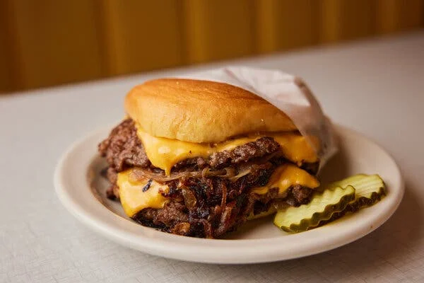 A double cheeseburger emerges from its wax wrapper. On the side are two crinkle-cut pickle slices.