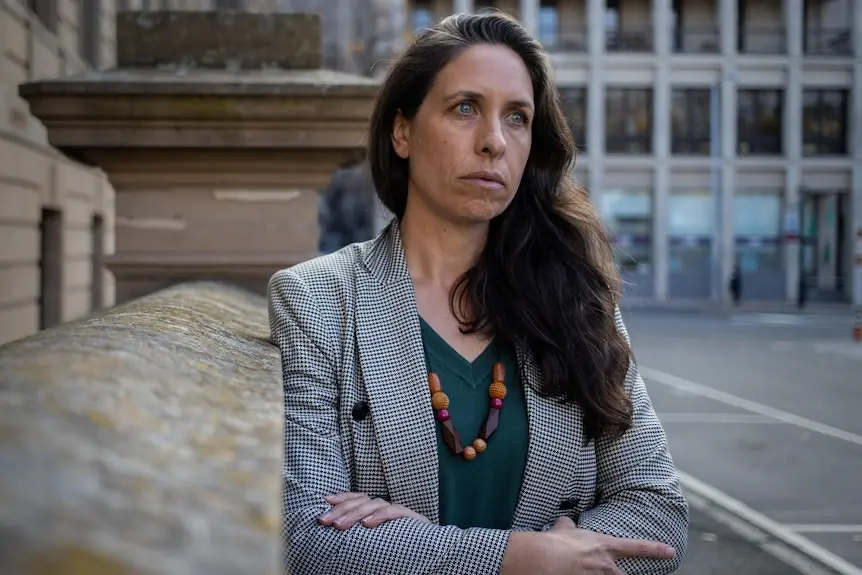 A woman leans against a sandstone building with her arms folded.