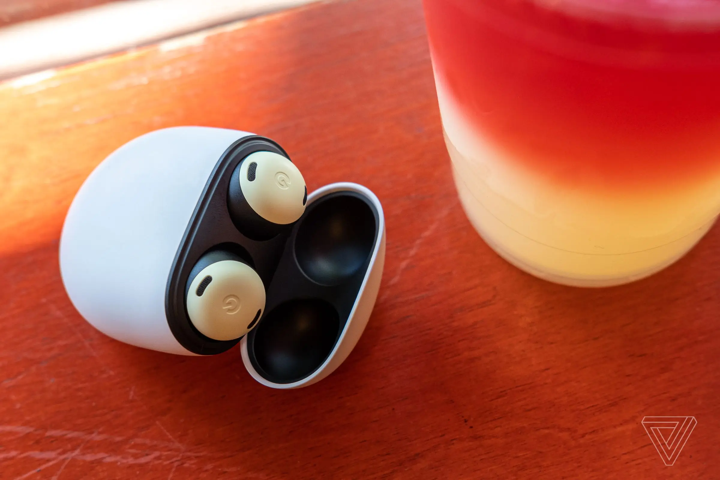 The yellow Pixel Buds Pro wireless earbuds sitting in their charging case with the lid open, resting on an orange table beside a plastic cup of colorful lemonade.