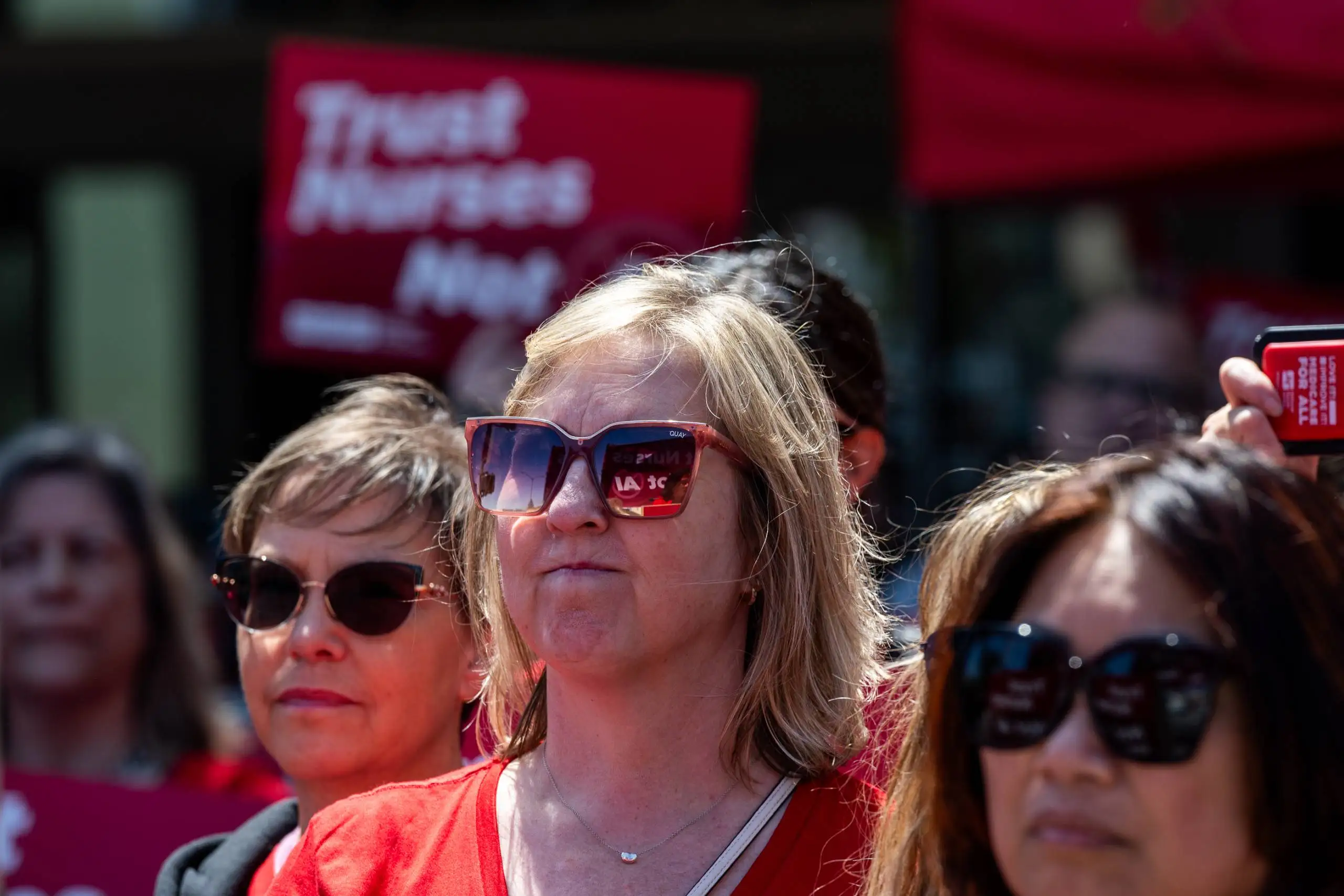 A woman wearing sun glasses and a red shirt stands in a crowd with red signs in the background.