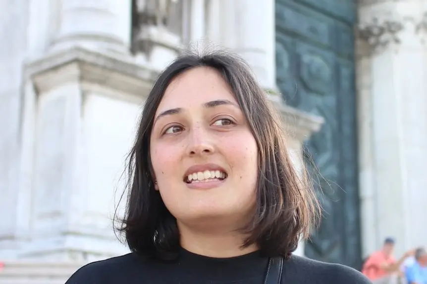 A young woman with a dark bob and black crew neck pictured in front of an old European looking building 