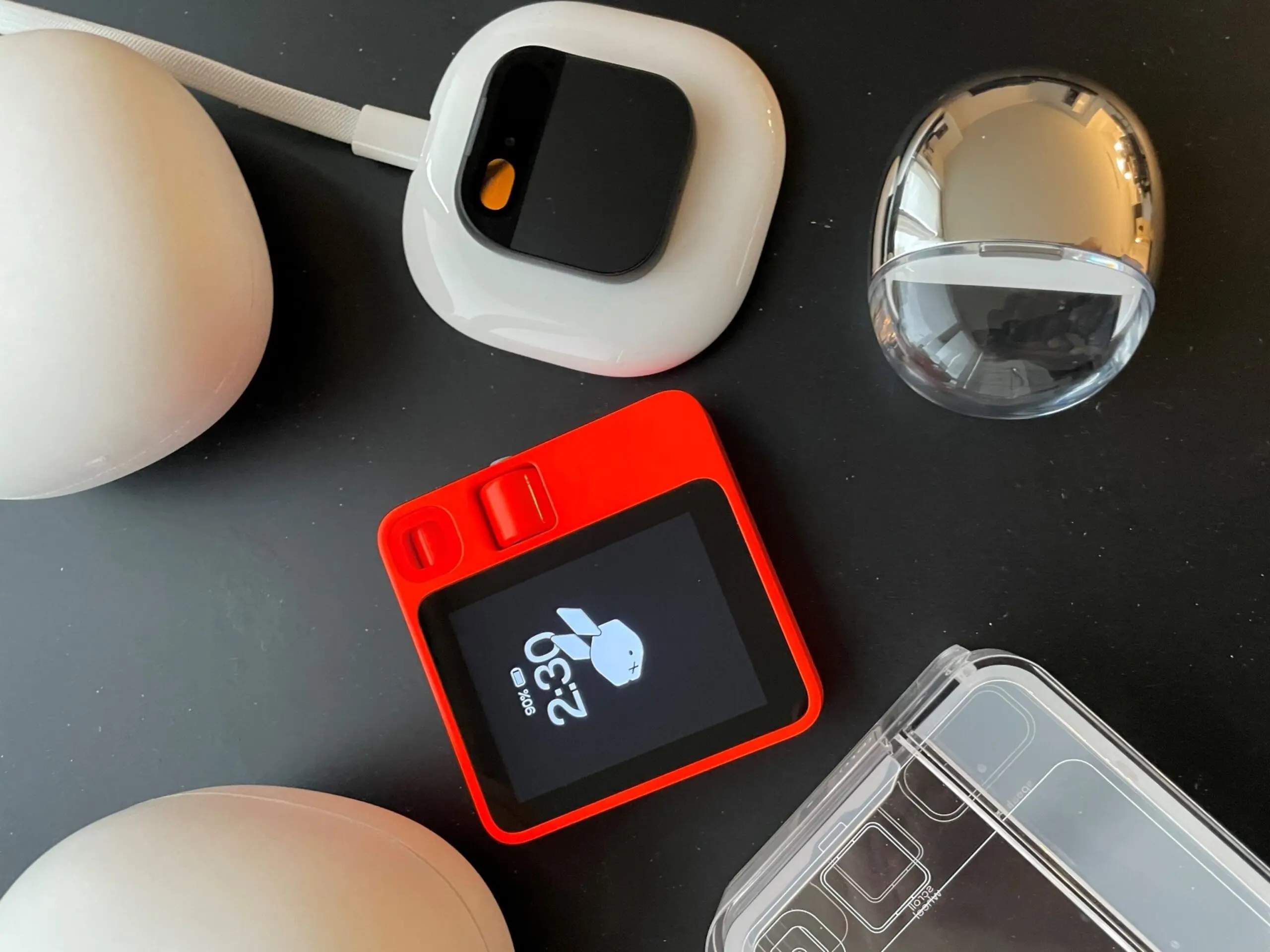 PHOTO: A Rabbit R1 and a Humane AI Pin are arranged on a table alongside their charging and carrying cases, April 29, 2024. 