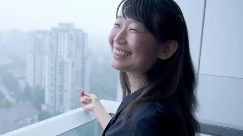 A woman with bangs smiles on a city balcony.