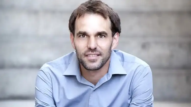 A man wearing a light blue dress shirt looks at the camera. He has brown hair and stubble.