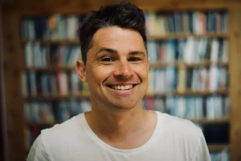 Michael Christie smiles widely into the camera with bookshelves in the background.